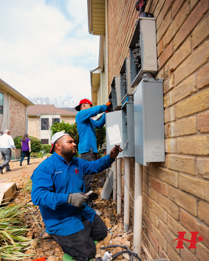 Two electricians from Powered By H installing outdoor electrical meter boxes on a brick building in Houston, TX.