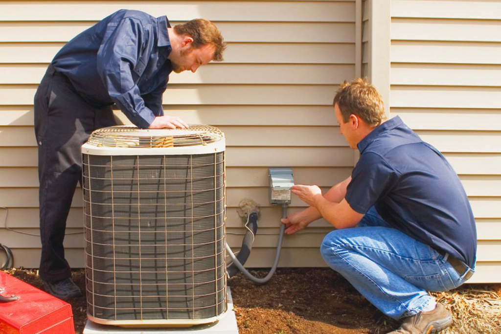 Two HVAC technicians performing maintenance or installation on an outdoor air conditioning unit for APM Heating and Air Conditioning in Columbia, MO.