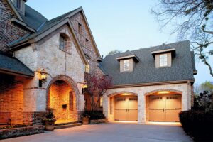 A stone and brick house featuring two light brown paneled garage doors with windows, illuminated at dusk by Overhead Door Company of Knoxville in Knoxville, TN