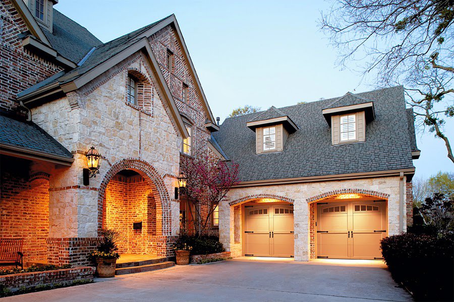 A stone and brick house featuring two light brown paneled garage doors with windows, illuminated at dusk by Overhead Door Company of Knoxville in Knoxville, TN