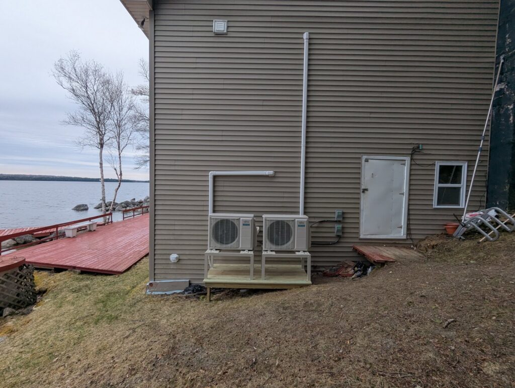 Two outdoor heat pump units installed on the side of a lakeside house by Kinney Family Heat Pumps in Augusta, ME.