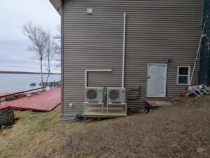 Two outdoor heat pump units installed on the side of a lakeside house by Kinney Family Heat Pumps in Augusta, ME.