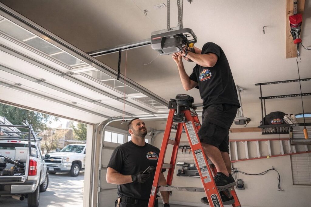 Two Baystate Garage Door technicians installing a garage door opener on a ladder in Milton, MA.