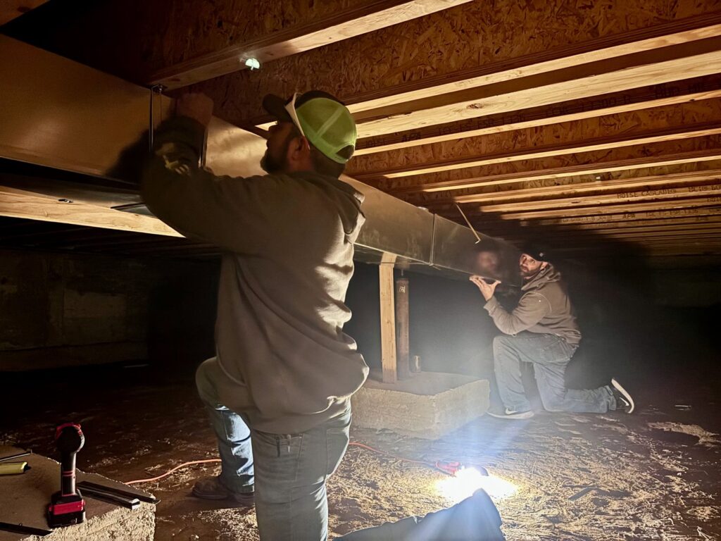 Two technicians from Lee's Heating & Cooling installing HVAC ductwork in a crawl space in Pueblo, CO.
