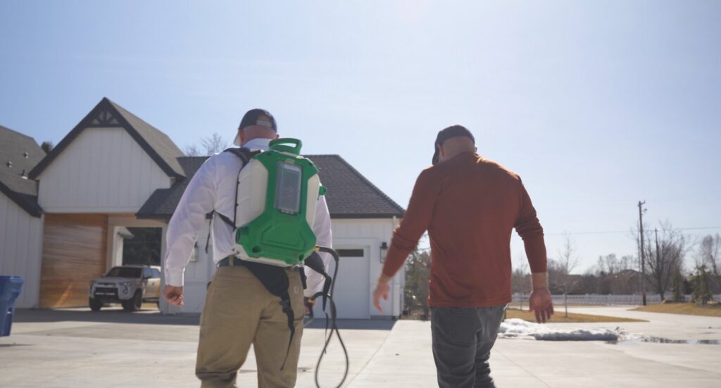 Two Hemi Pest Control technicians, one with a backpack sprayer, walking towards a house for a service in Meridian, ID.