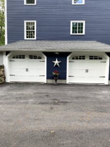 Two white garage doors with decorative hardware and top windows installed on a blue house by Snowbird Garage Doors Company in Worcester, MA.
