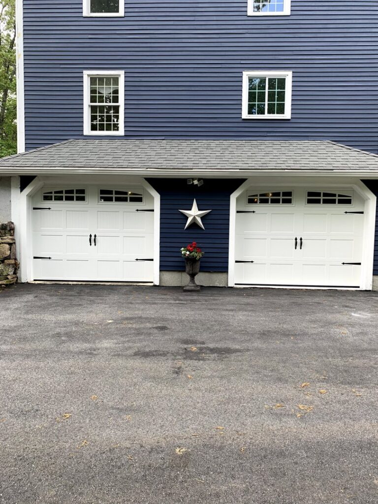 Two white garage doors with decorative hardware and top windows installed on a blue house by Snowbird Garage Doors Company in Worcester, MA.