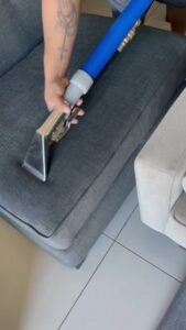 A professional cleaning a grey sofa cushion with an upholstery cleaning tool, performed by Good Guys Carpet Cleaning in Katy, TX.