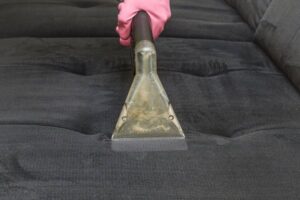 A gloved hand using a professional upholstery cleaning tool on a dark fabric sofa by Mid State Contract Cleaners in Nashville, TN.