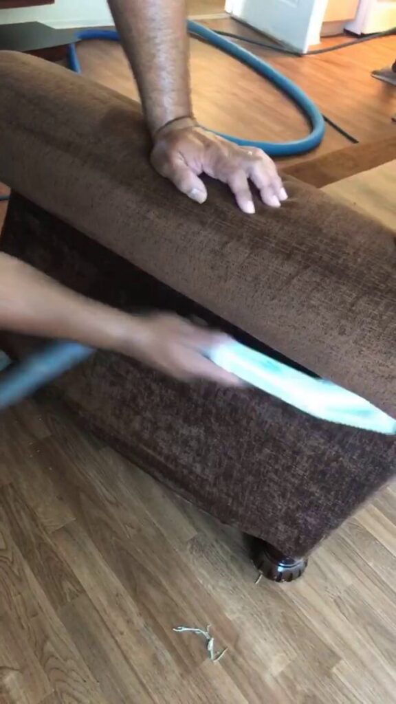 A professional cleaning a brown upholstered sofa with a specialized tool at Alex's Janitorial and Carpet Cleaning in Bakersfield, CA