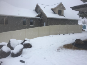 A light-colored vinyl fence installed on a snowy hillside next to a house by The Best Fence Company in Spokane, WA.