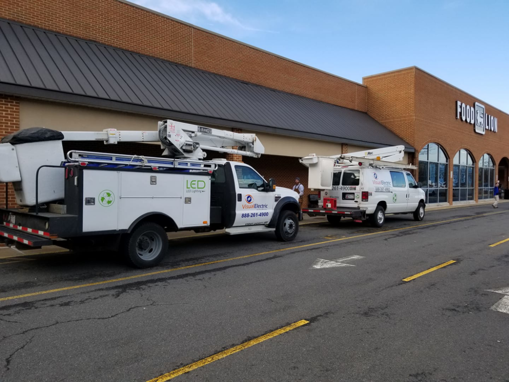 Two Visual Electric, Inc. service vehicles, including a bucket truck for LED lighting, parked outside a Food Lion in Frederick, MD.