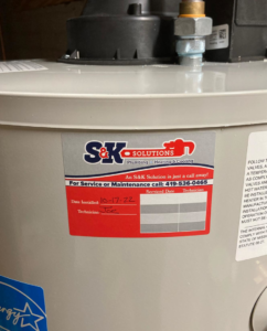 A close-up of a water heater with a service sticker from S & K Solutions, showing installation date in Petersburg, MI