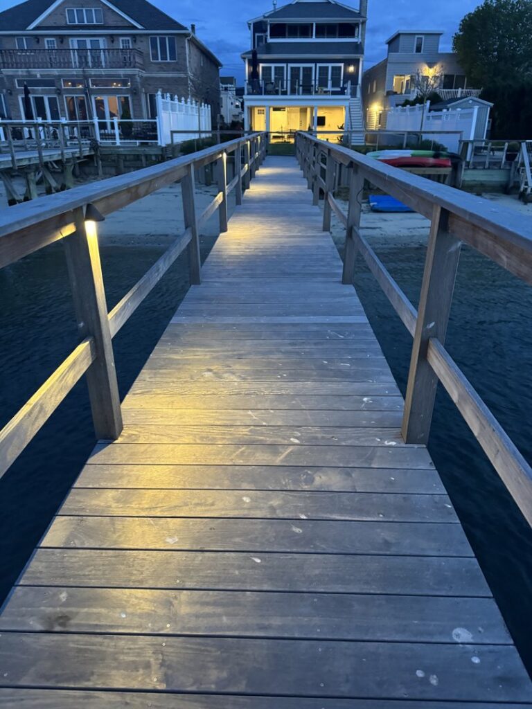 A wooden pathway on a dock with installed lighting leading to waterfront houses, completed by New York Electrical Design in Freeport, NY