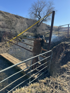 A worker welding a metal fence post over a stream during a fencing project by Deer Creek Ag Services in Ranchester, WY.
