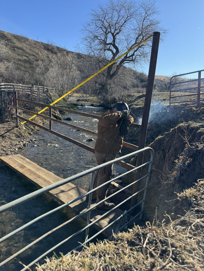 A worker welding a metal fence post over a stream during a fencing project by Deer Creek Ag Services in Ranchester, WY.