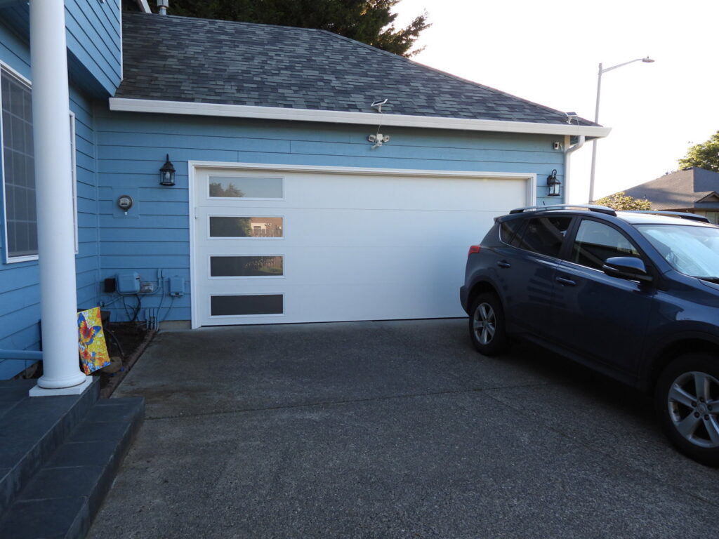 A white modern garage door with vertical windows on a blue house, installed by Coast To Coast Garage Door LLC in Vancouver, WA.