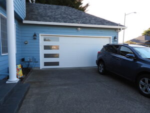 A white modern garage door with vertical windows on a blue house, installed by Coast To Coast Garage Door LLC in Vancouver, WA.
