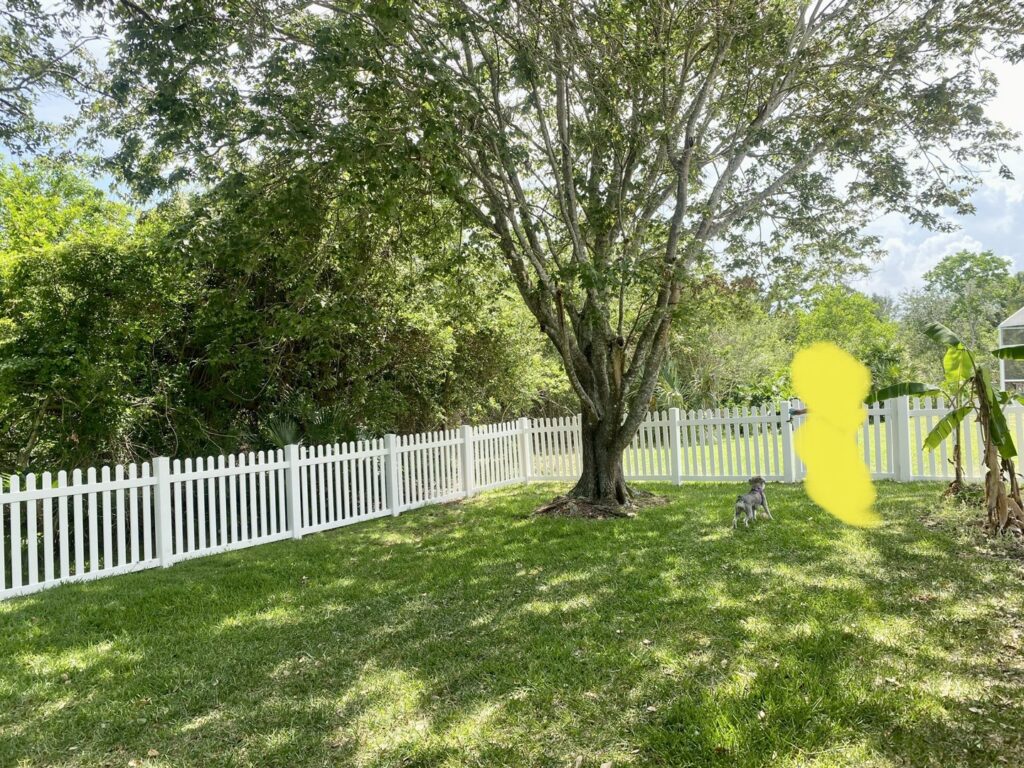 A charming white picket fence enclosing a lush green backyard, perfect for pets, installed by Exacta FENCE LLC in Jacksonville, FL.