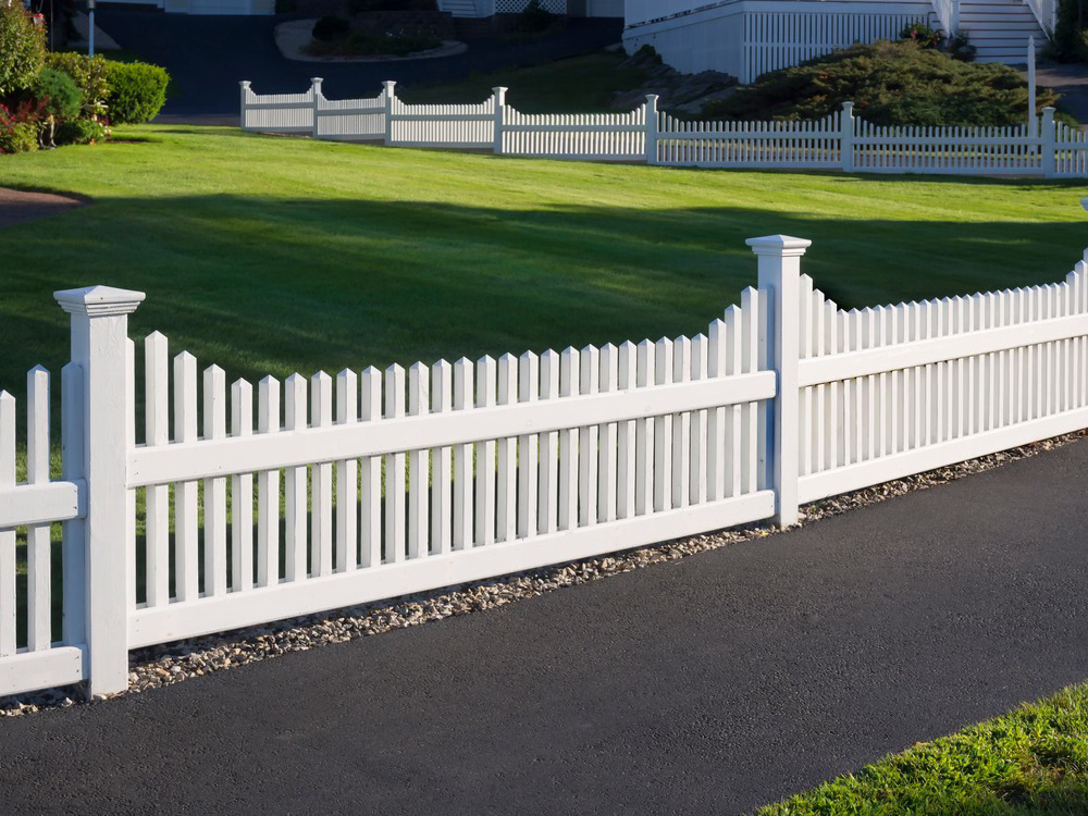 A classic white picket fence installed along a residential driveway and green lawn by New Bedford Fence in New Bedford, MA.