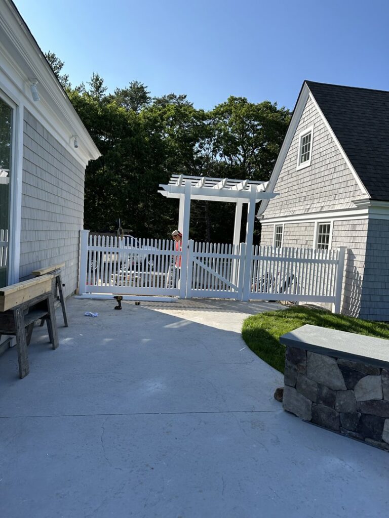 View of a white picket fence with a gate and arbor from a residential patio, installed by New England landscaping and fence inc in Lynn, MA.