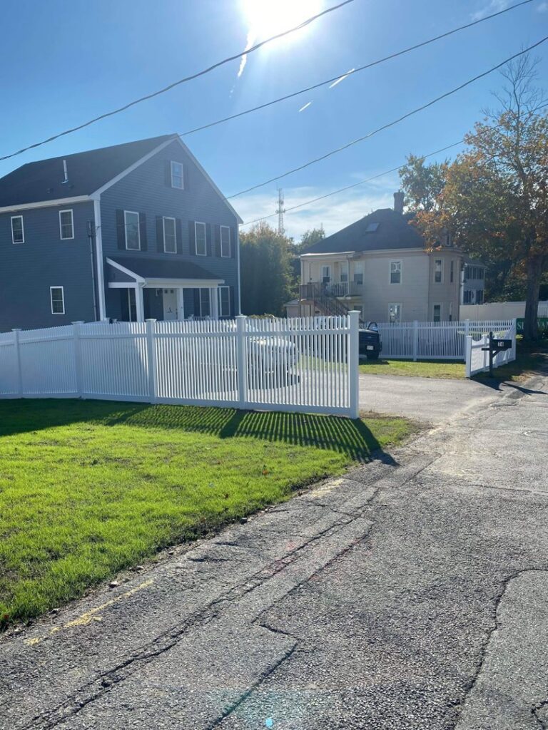 A long white picket fence with a gate installed along a residential street by New England landscaping and fence inc in Lynn, MA.