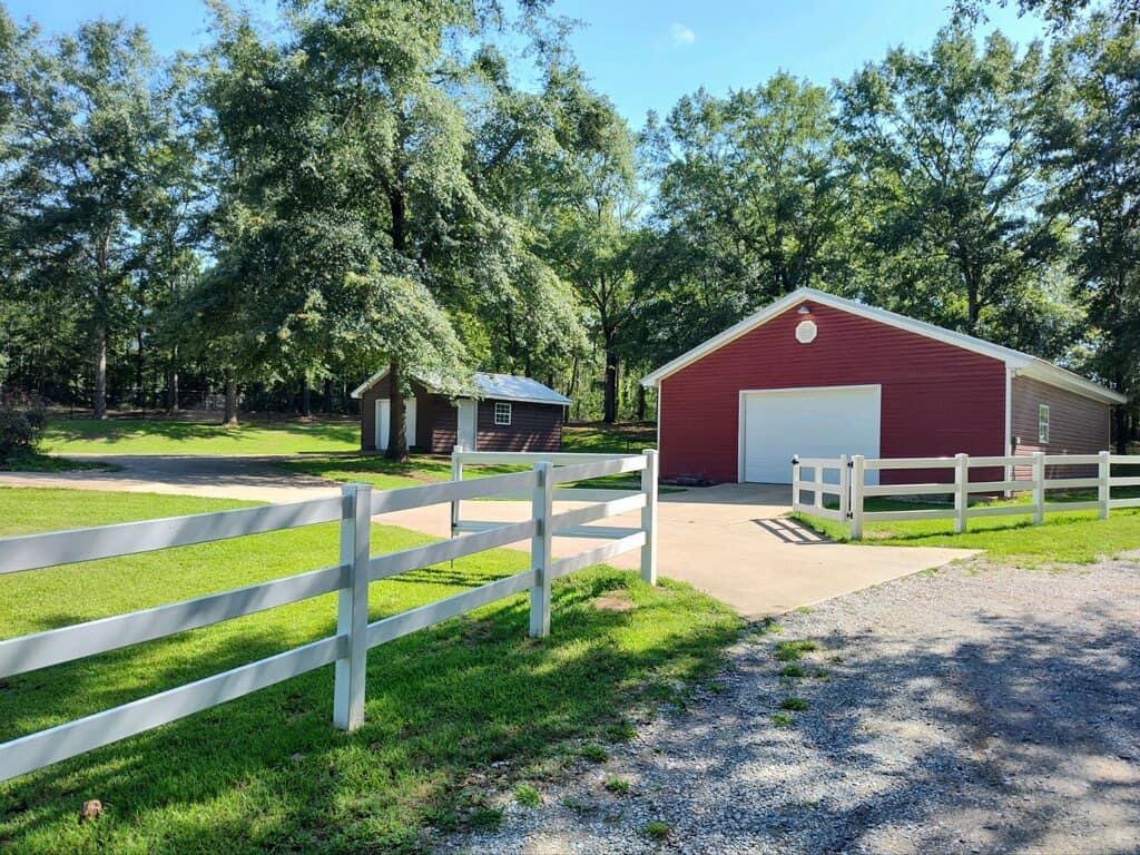 A white post-and-rail fence installed in front of a red barn by TC Fencing LLC in Tuscaloosa, AL.