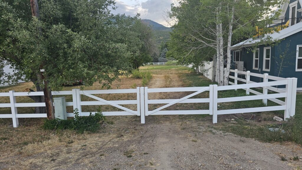 A white three-rail vinyl farm fence with a gate along a dirt path, installed by All American Vinyl in Provo, UT.