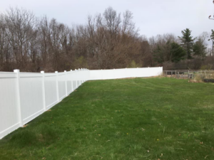 An extensive white vinyl fence installed along the edge of a green lawn, bordering a wooded area by David Platania Fence in Derry, NH.