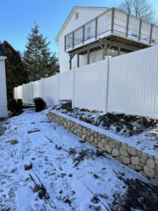 A newly installed white vinyl privacy fence by Hunter & Sons Fence in Burlington, MA, with a stone wall and snow.