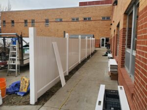 A white vinyl fence installation in progress next to a brick building by Pride Fence in Peabody, MA.