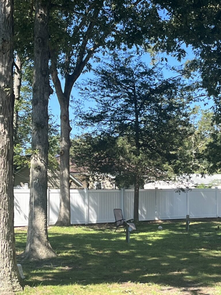 A white vinyl privacy fence visible through trees in a residential backyard by MAC Fencing LLC in Toms River, NJ