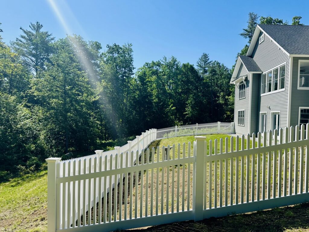 A white vinyl picket fence installed on a sloped residential yard by Ultra Fence LLC in Brookline, NH.