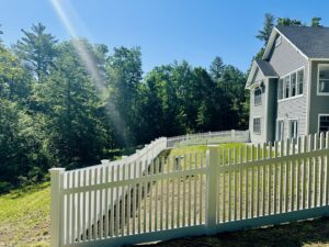 A white vinyl picket fence installed on a sloped residential yard by Ultra Fence LLC in Brookline, NH.