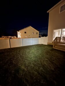 A newly installed white vinyl privacy fence in a backyard at night, illuminated by a light, by Vision One Construction in Schenectady, NY