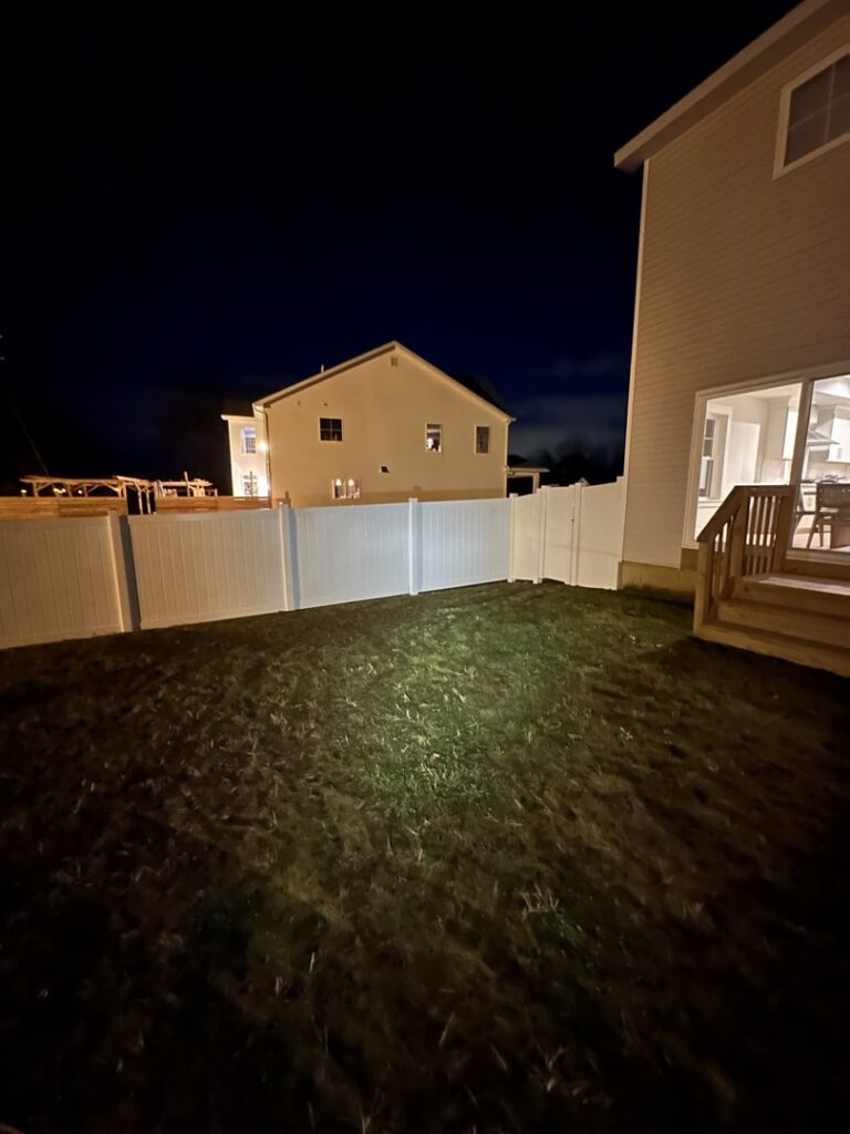 A newly installed white vinyl privacy fence in a backyard at night, illuminated by a light, by Vision One Construction in Schenectady, NY