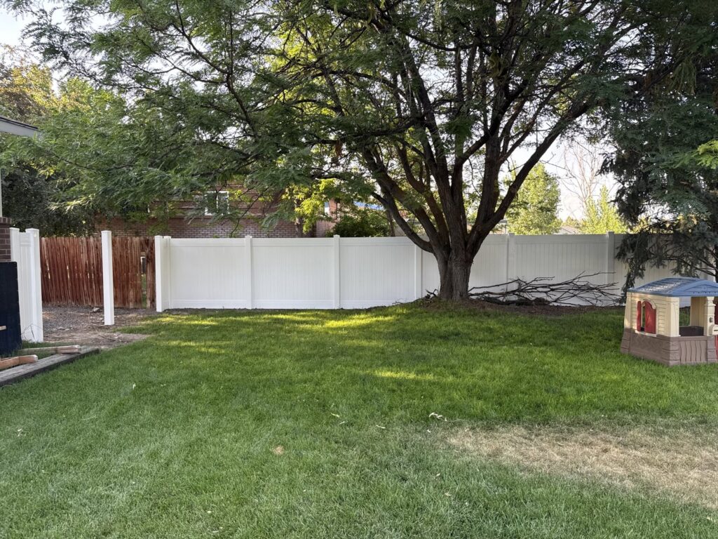 A white vinyl privacy fence installed in a backyard, partially shaded by a large tree, by Sublime Fencing LLC in North Logan, UT.