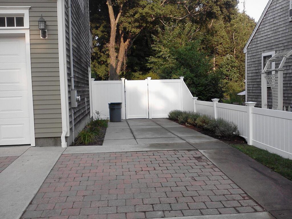 A white vinyl privacy fence with a gate installed next to a garage and driveway by OMA Fence,LLC in Weymouth, MA
