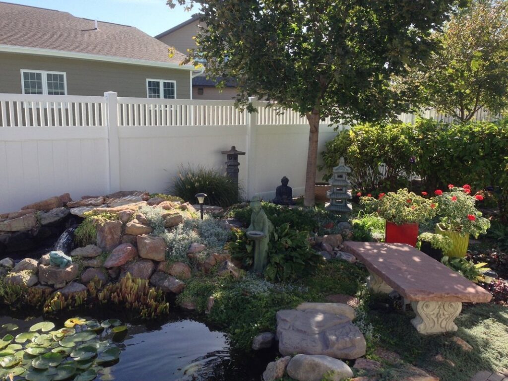 A white vinyl privacy fence installed around a decorative garden and pond area by Schumacher Fencing, LLC in Columbus, NE.
