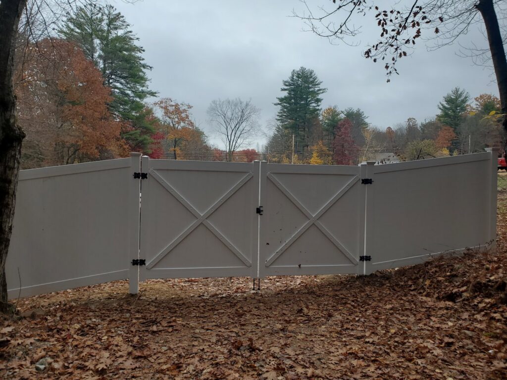 A white vinyl privacy fence with a decorative gate installed by MH Fence in Weare, NH, in a wooded setting.