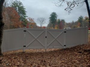 A white vinyl privacy fence with a decorative gate installed by MH Fence in Weare, NH, in a wooded setting.