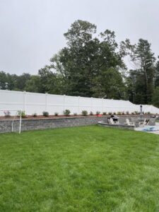 A white vinyl privacy fence installed along a retaining wall with plants by OMA Fence,LLC in Weymouth, MA