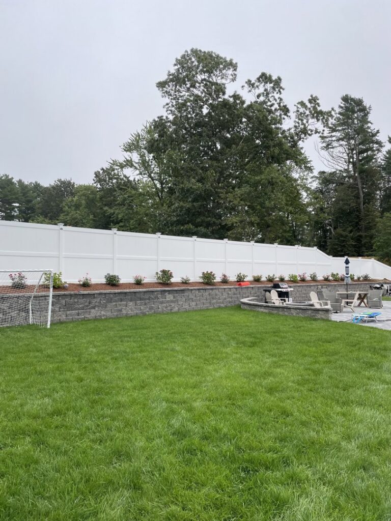 A white vinyl privacy fence installed along a retaining wall with plants by OMA Fence,LLC in Weymouth, MA