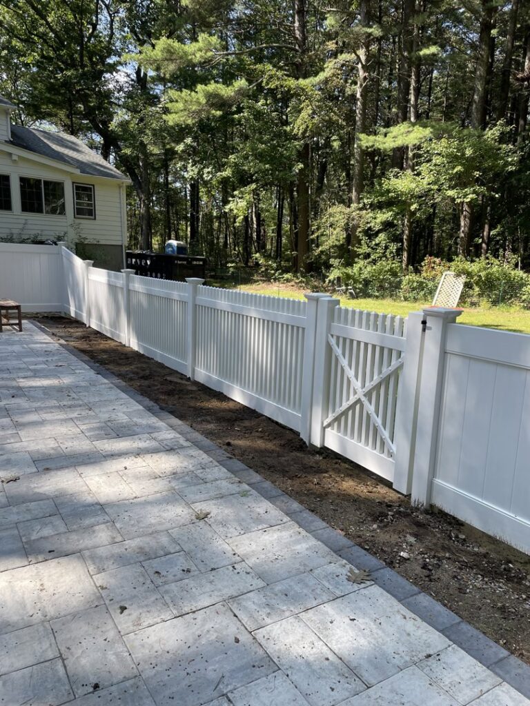 A newly installed white vinyl privacy fence with a gate next to a patio by OMA Fence,LLC in Weymouth, MA