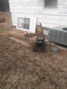 A humane wildlife trap set near a house foundation and AC unit, demonstrating wildlife control by Bug Busters LLC in Norman, OK.