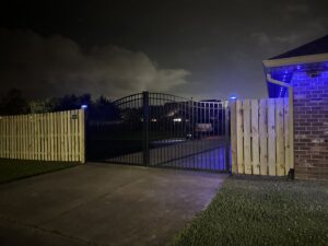 A wood fence with a black metal gate illuminated by blue lights at night, installed by Southern Fencing, LLC in Houma, LA.