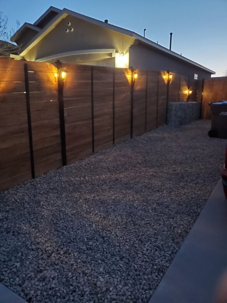 A newly installed wood fence on a stone wall illuminated by solar lights at dusk, completed by MJ Custom Fencing in Las Cruces, NM.