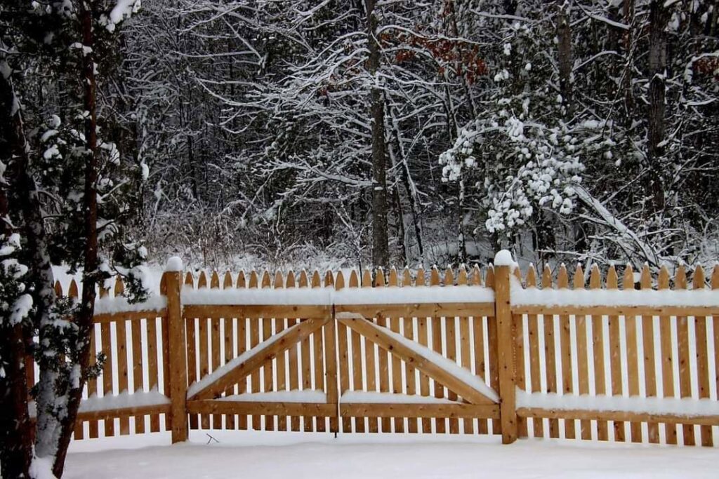 A charming wooden picket fence and gate covered in a fresh blanket of snow, installed by Sullivan & Sons Fence LLC in Hudson, NH.