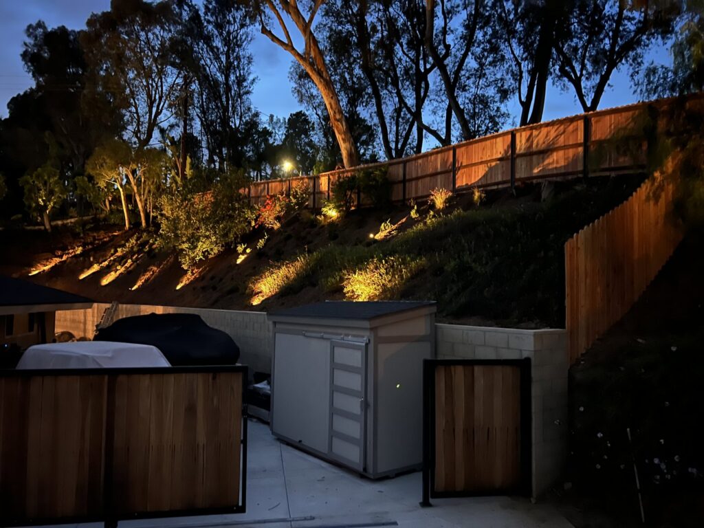A wood privacy fence installed on a sloped backyard property, illuminated by landscape lighting by True American Fence Supply and Construction in Anaheim, CA.