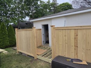 A wooden fence under construction next to a building, with deck framing visible, by Homestead Fence in Cumberland, RI.